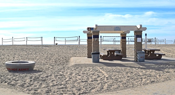 Newland Ramada A sits on a paved path (wheelchair accessible). There are two picnic tables and a fire ring adjacent to the area. Volleyball courts line the sand several yards behind the picnic area.